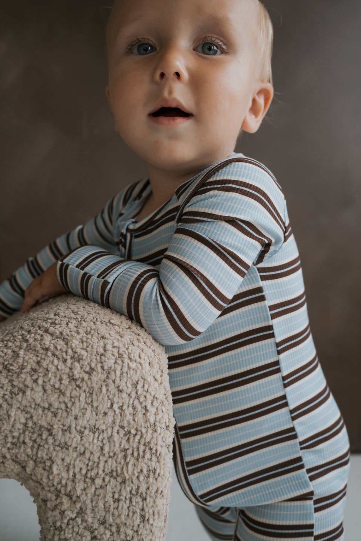 A young child wearing blue and brown striped ribbed Bamboo Zip Pajamas | Dean leans on a textured surface.