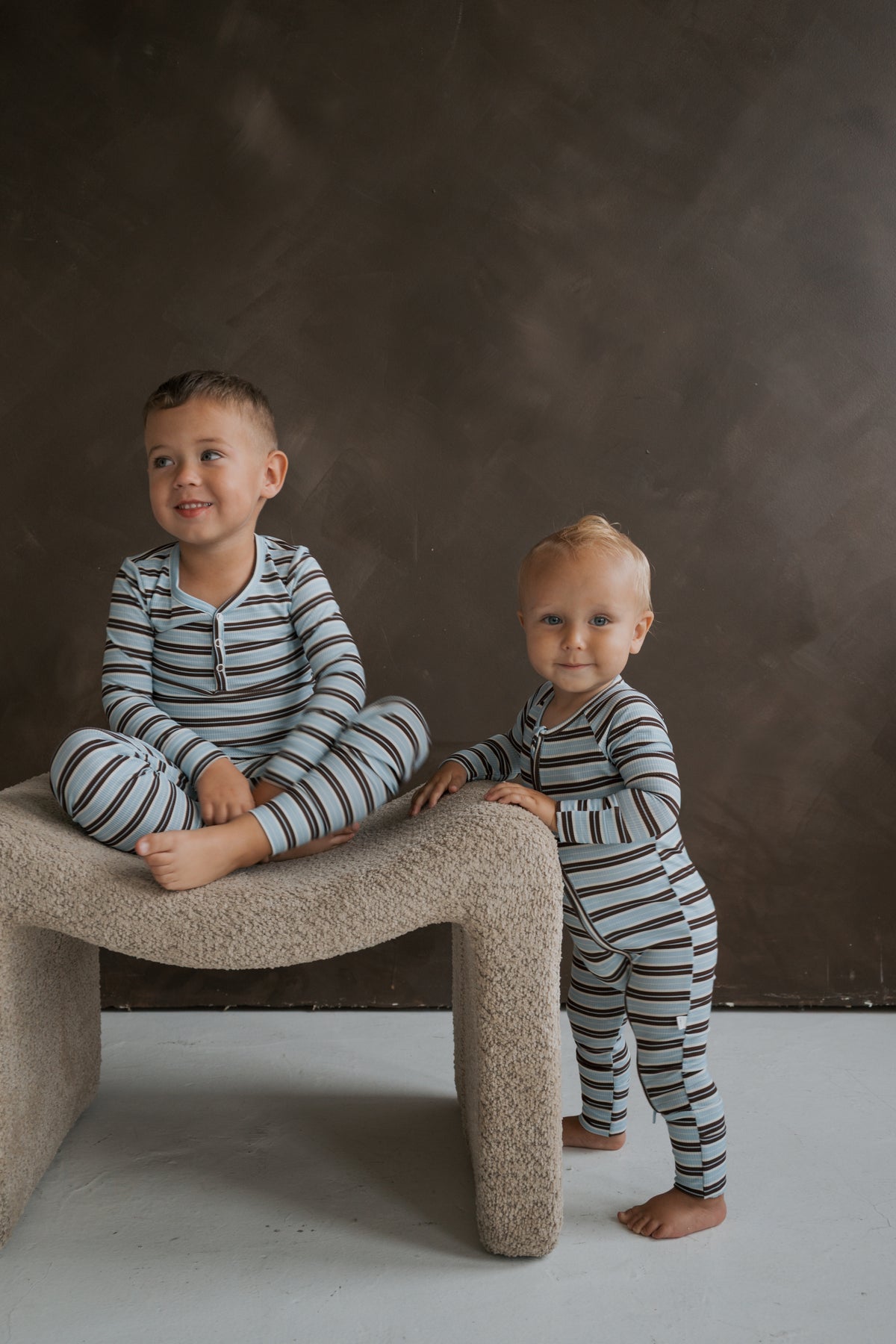 Two young children wearing matching blue and brown striped Bamboo Two Piece Pajamas | Dean, with one child sitting on a stool and the other standing beside it.