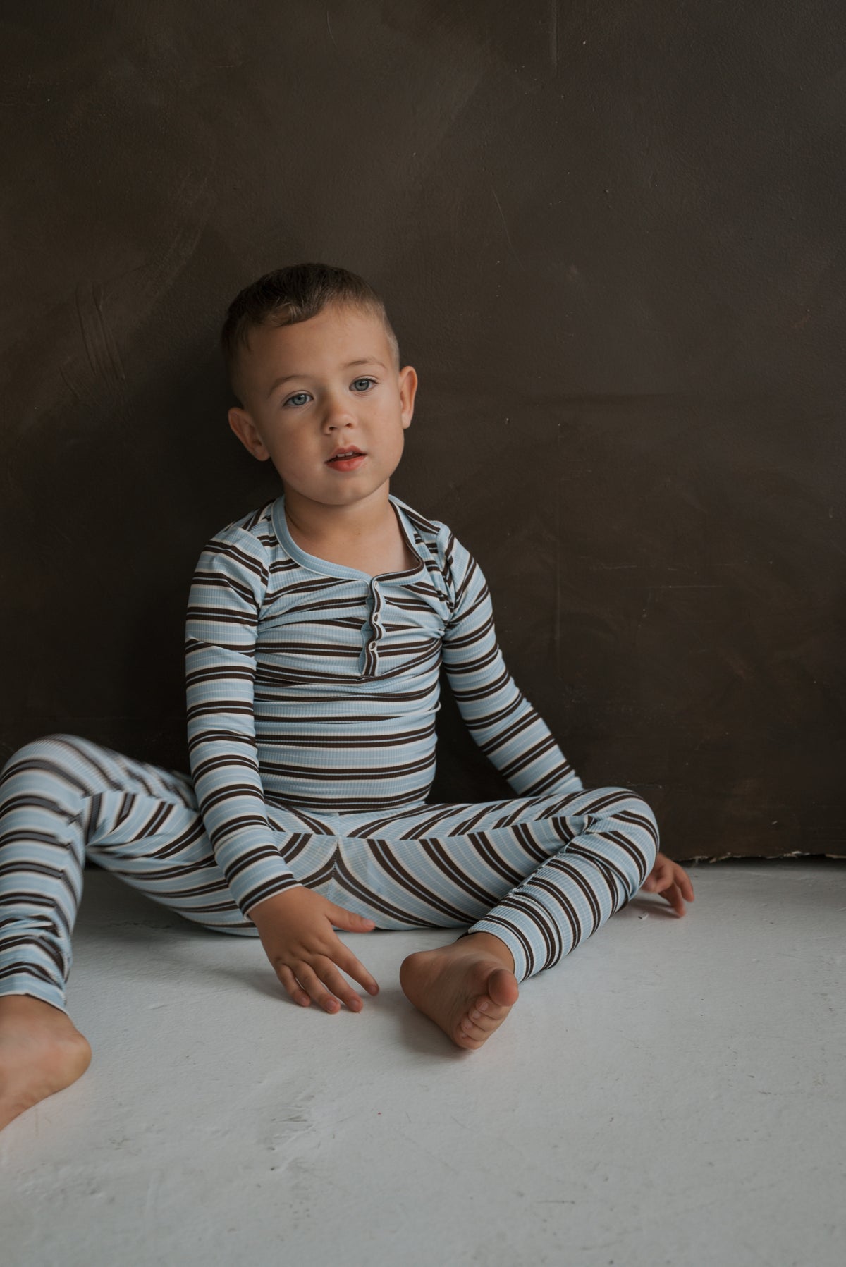A young boy sitting on the floor wearing Bamboo Two Piece Pajamas | Dean in a long-sleeved shirt and pants with blue and brown horizontal stripes.