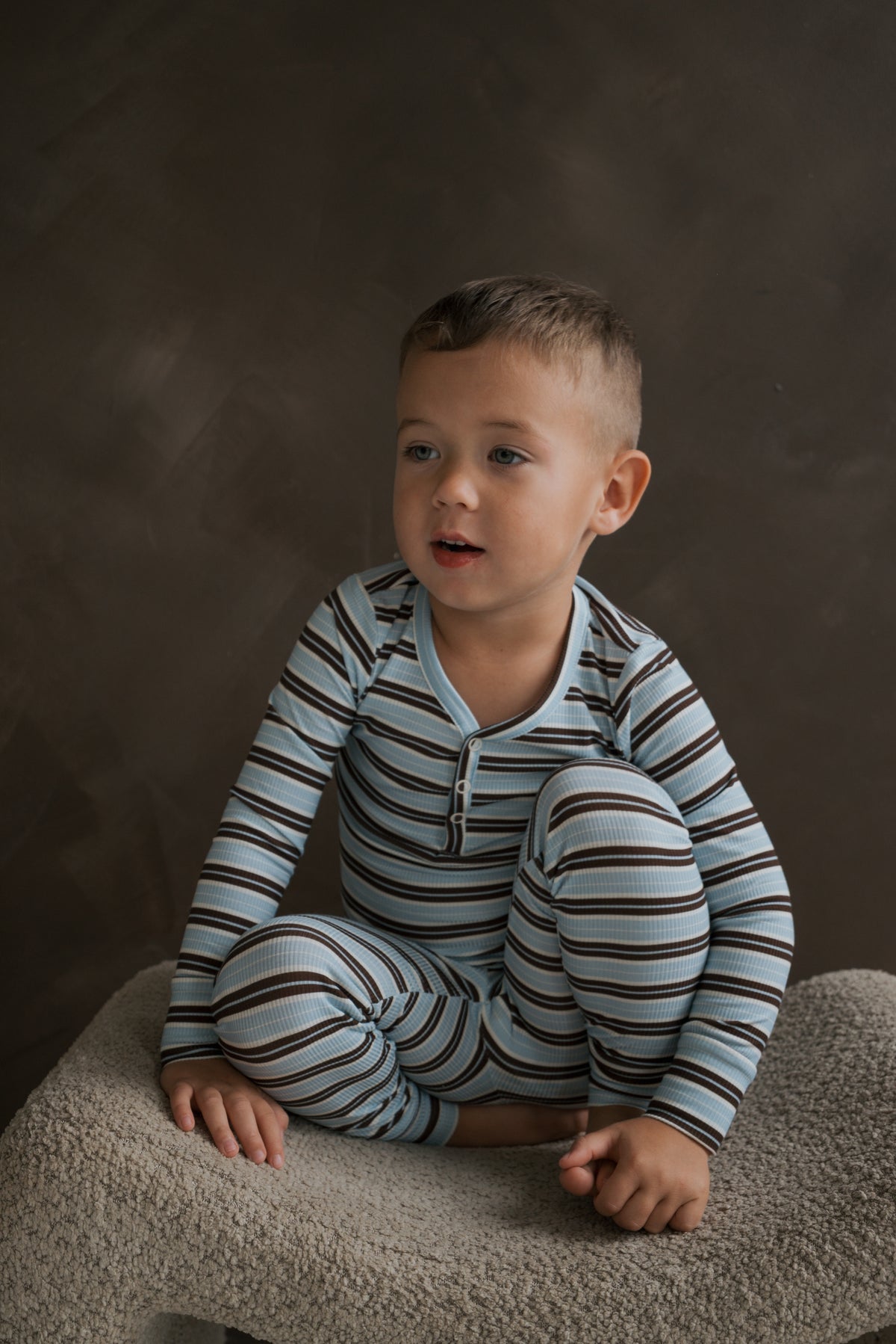 A young boy wearing the blue and brown striped Bamboo Two Piece Pajamas | Dean by forever french baby, featuring a ribbed long-sleeved top with a henley neckline and matching bottoms.