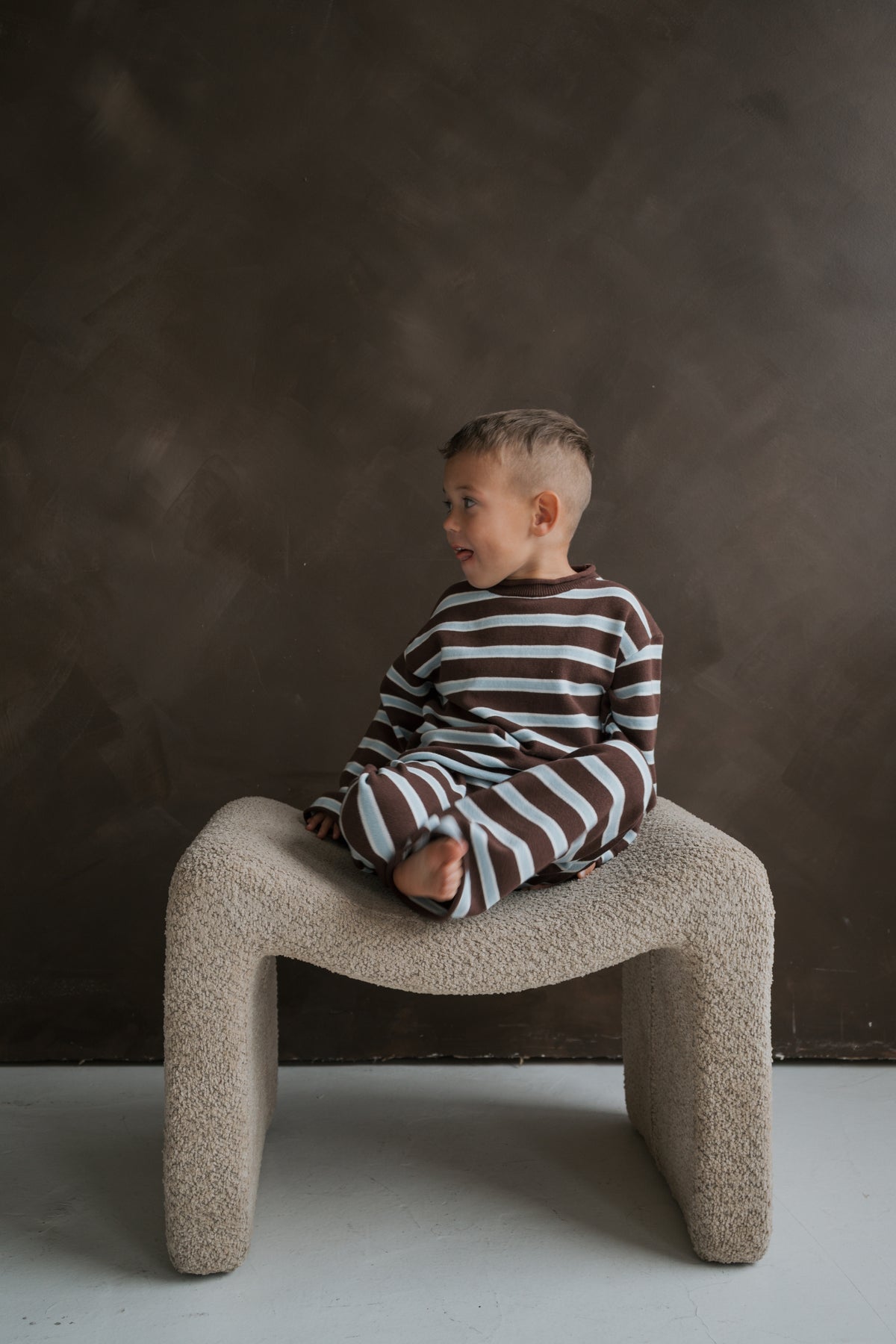 A young child sits cross-legged on a textured stool wearing a Knit Pant Set | Garrett *PRE-ORDER* featuring a 100% cotton long-sleeved shirt and pants with brown and light blue horizontal stripes.
