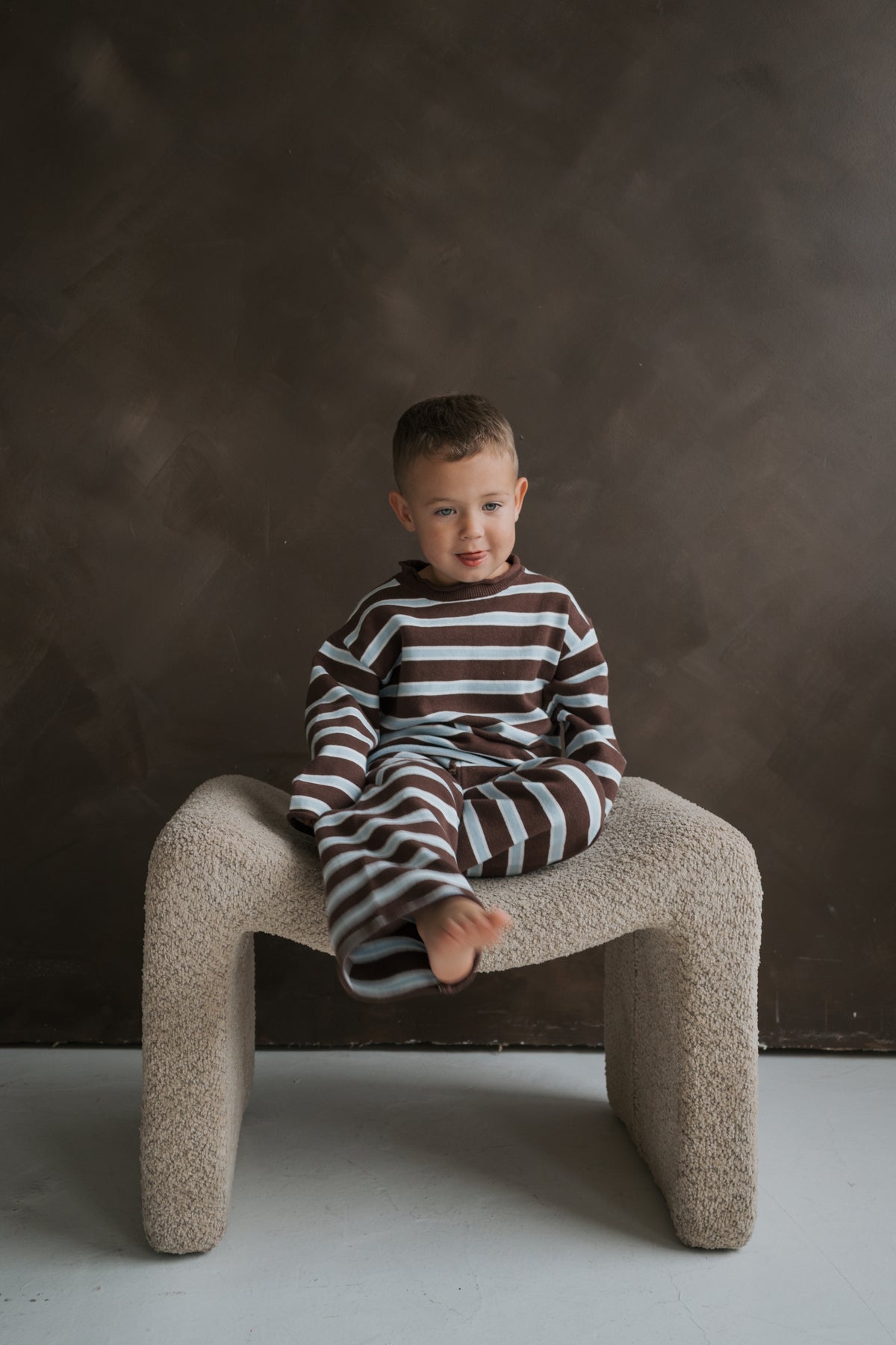 A young child wears a forever french baby Knit Pant Set in a brown and blue striped knit as they sit on a textured stool.