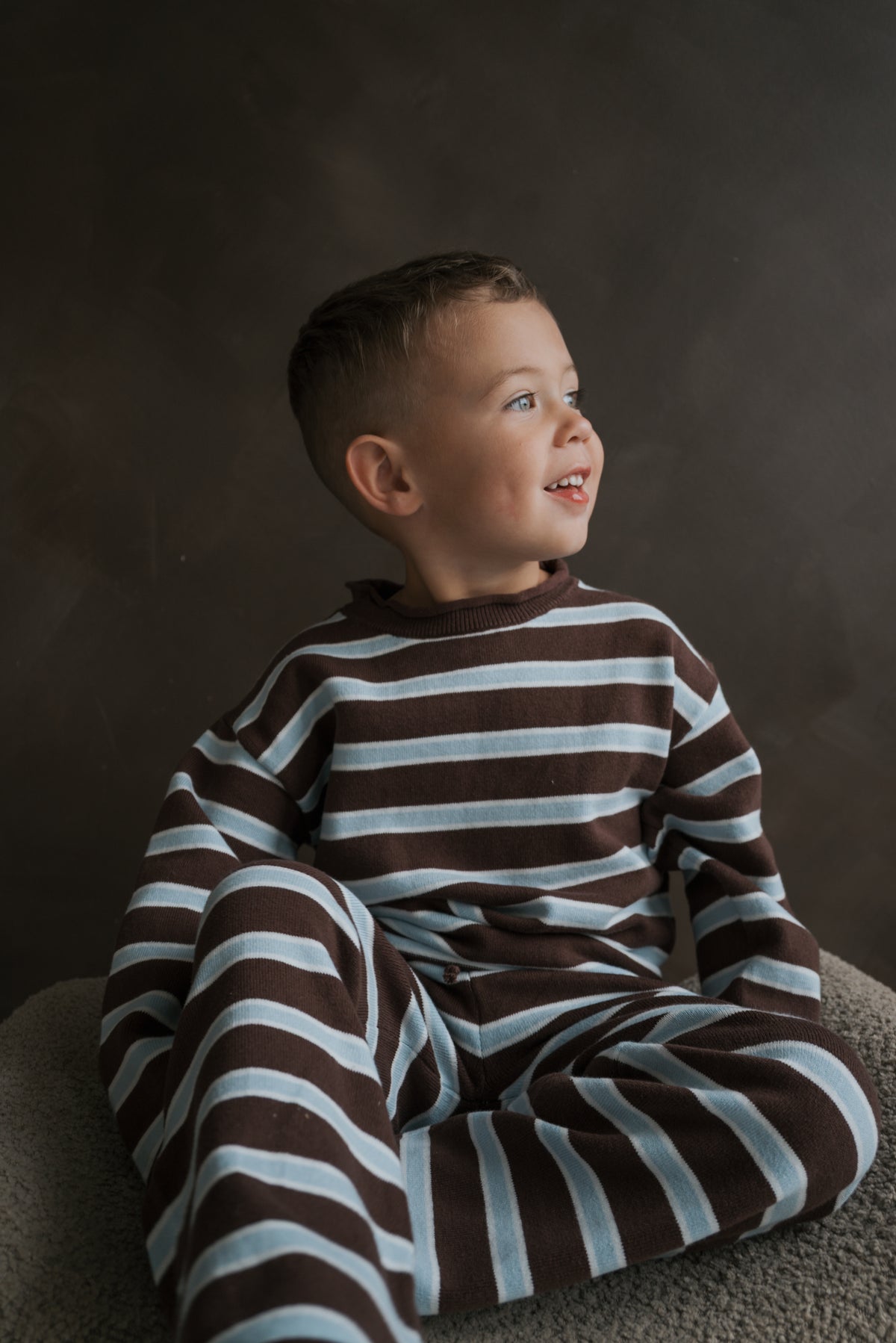 A young boy wears the forever french baby Knit Pant Set | Garrett *PRE-ORDER*, a 100% cotton set in brown and light blue stripes featuring a long-sleeved shirt and matching pants.