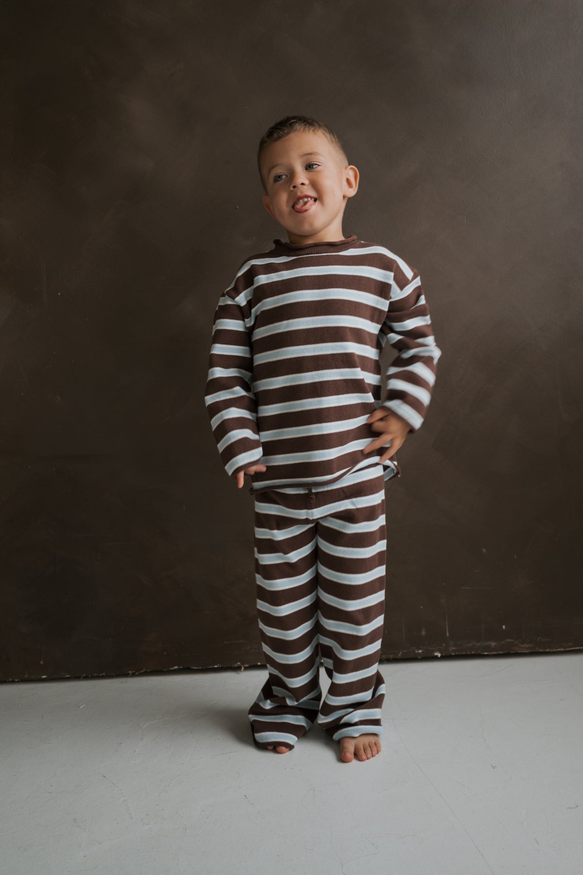 A young child wearing a Knit Pant Set | Garrett by forever french baby, featuring a long-sleeved shirt and matching pants with horizontal brown and light blue stripes, sticking their tongue out.