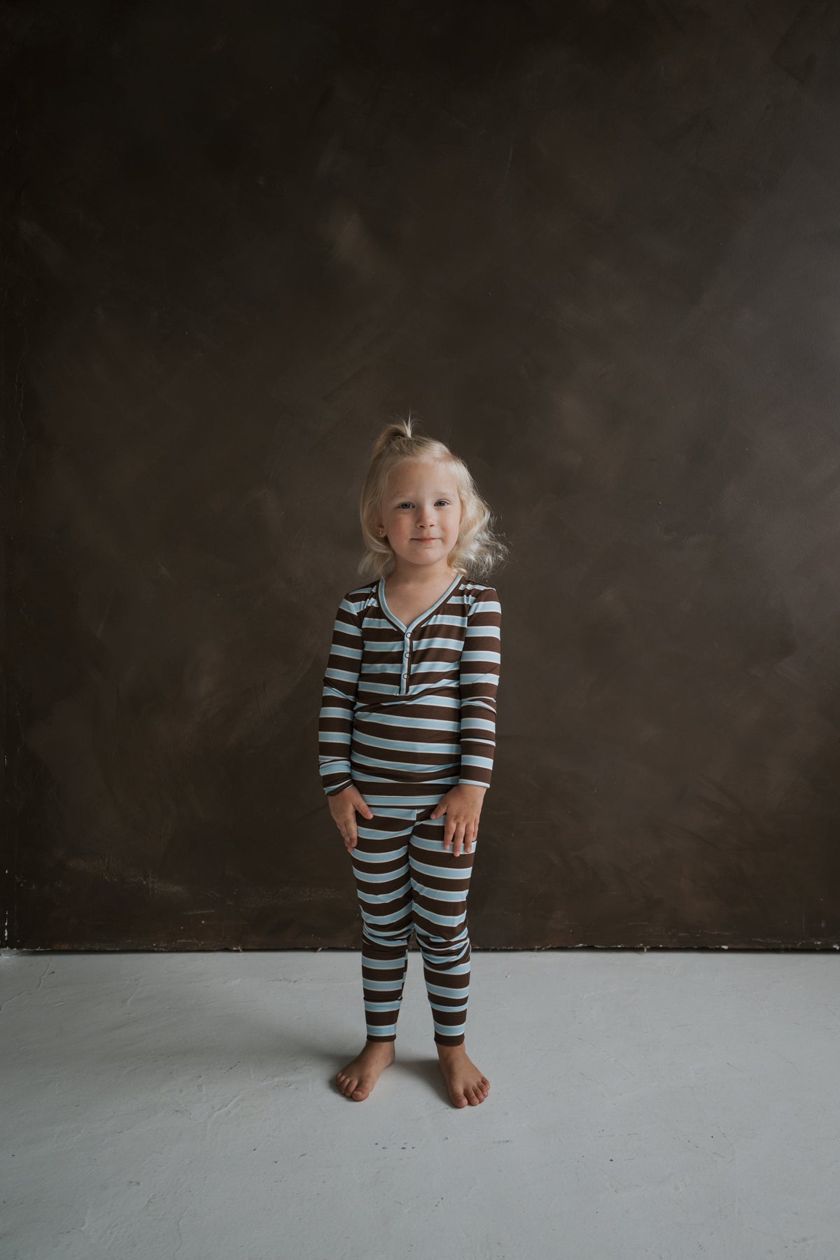A child in Bamboo Two Piece Pajamas | Garrett, a breathable henley-style pajama top and matching leggings with brown and light blue horizontal stripes.