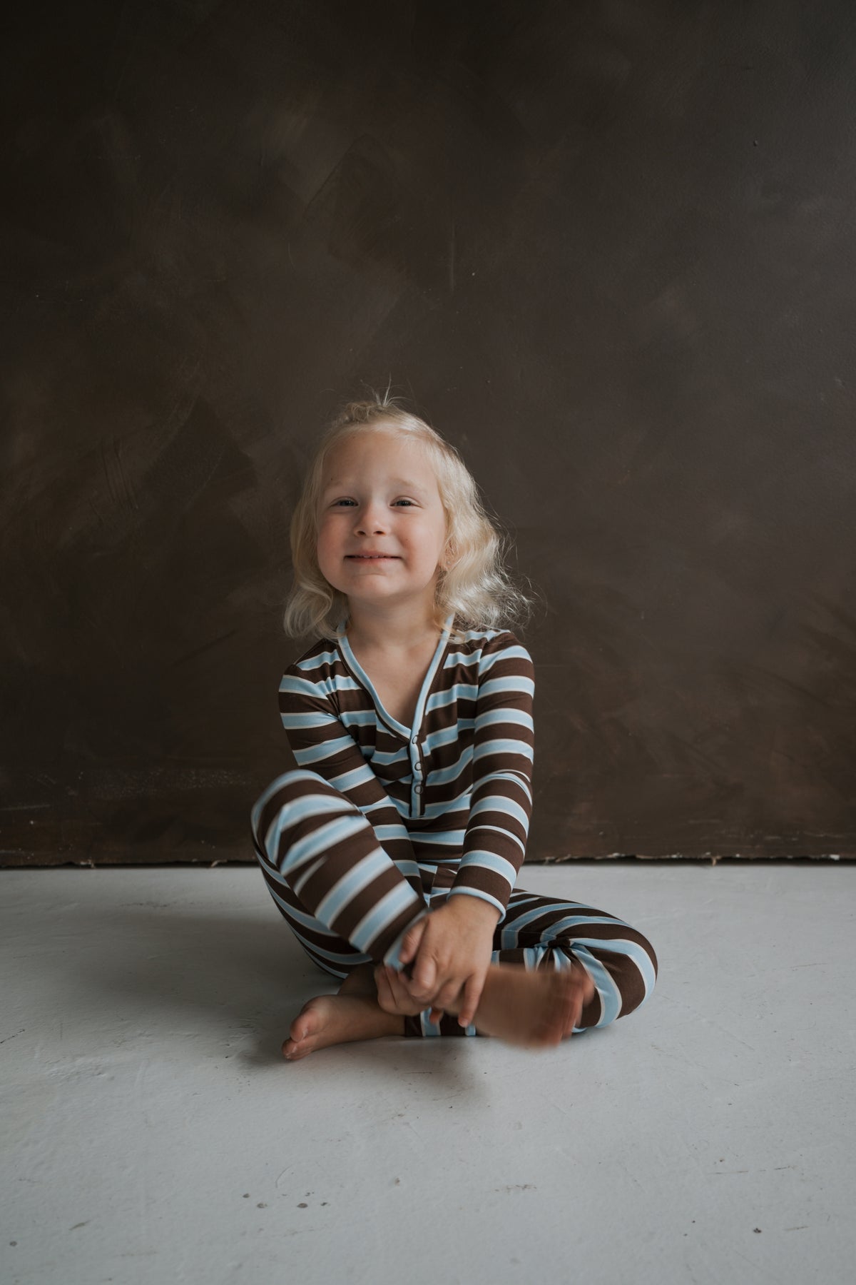A child sits cross-legged wearing Bamboo Two Piece Pajamas | Garrett, which have brown and light blue horizontal stripes.