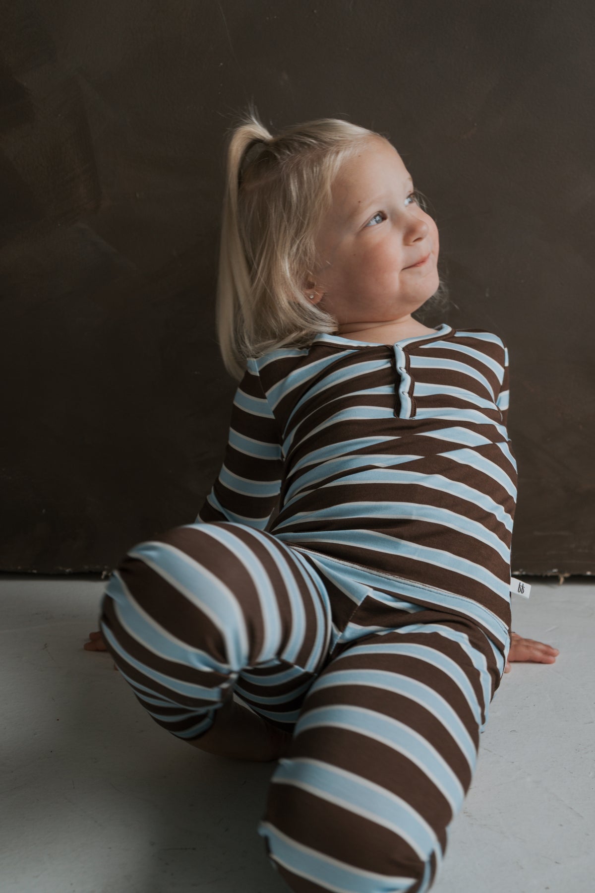 A young child sits on the floor wearing brown and light blue striped Bamboo Two Piece Pajamas | Garrett by forever french baby.