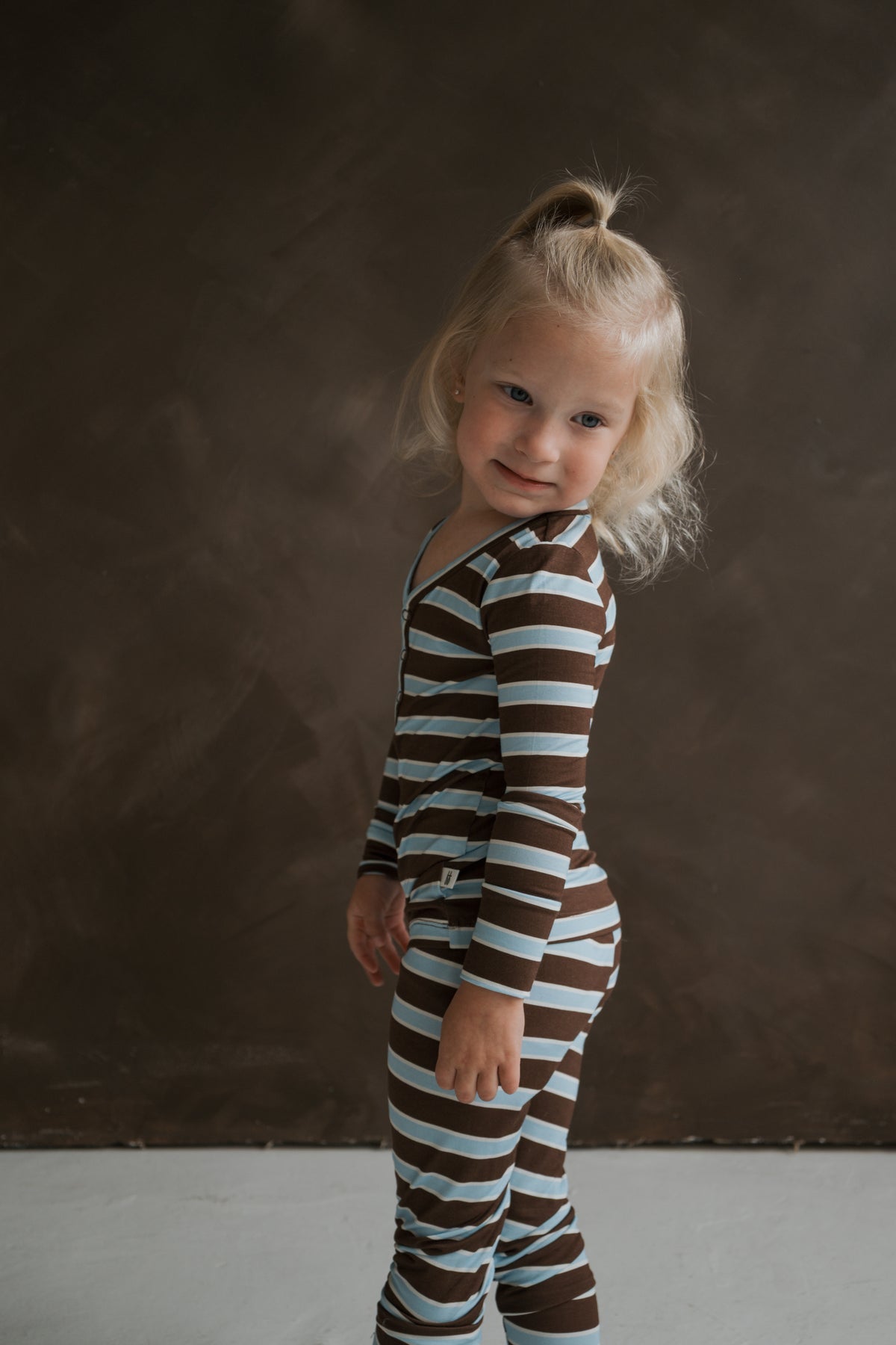A toddler wearing forever french baby Bamboo Two Piece Pajamas | Garrett, featuring a long-sleeved shirt and leggings with brown and light blue horizontal stripes.