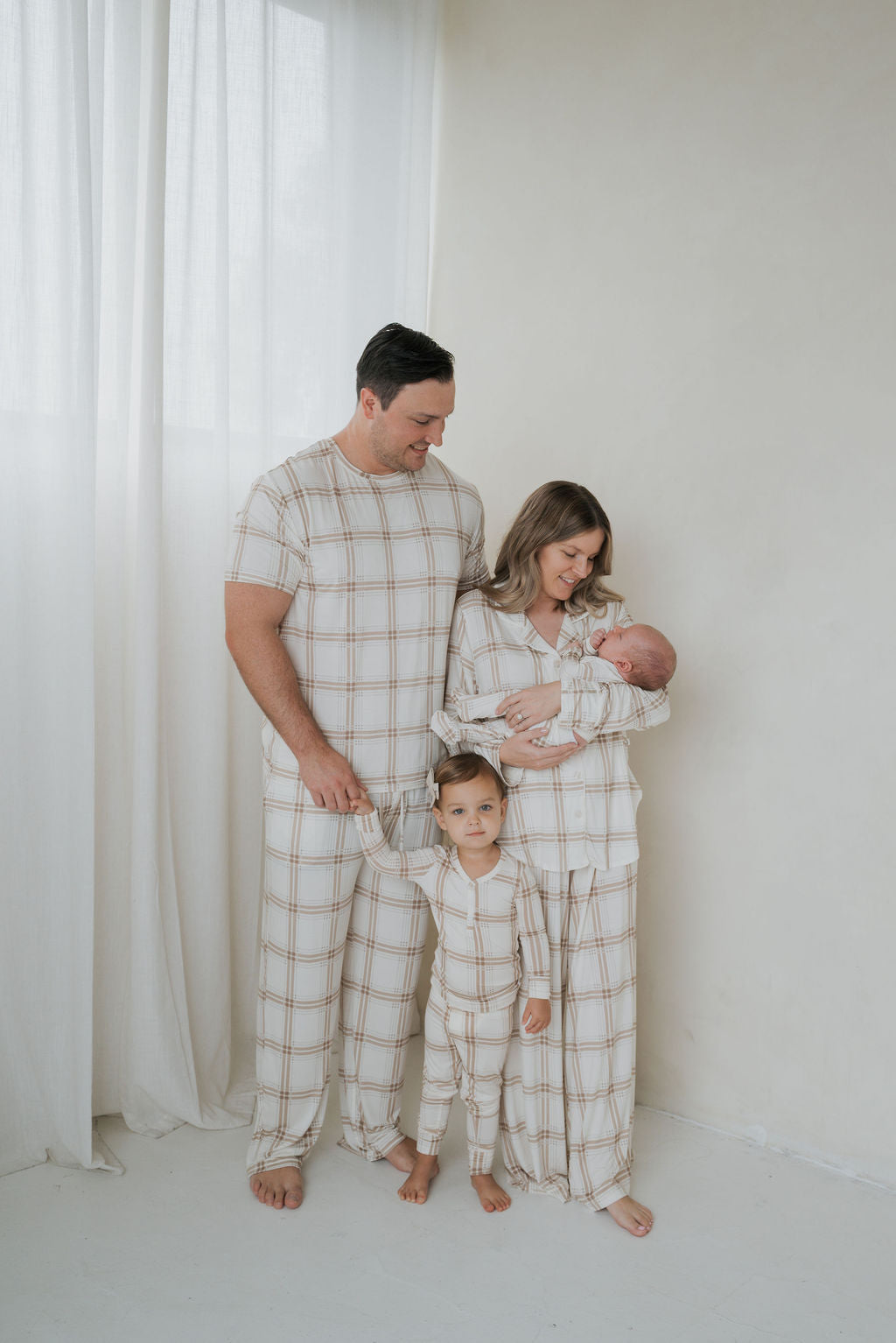 A family of four wearing matching tan and white Bamboo Two Piece Pajamas | Ellis Plaid, with the parents holding their two young children.