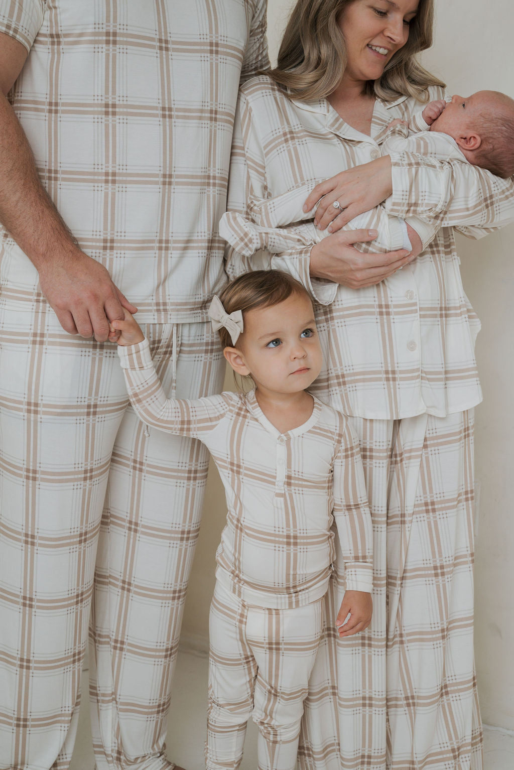 A family of four wears matching Bamboo Two Piece Pajamas | Ellis Plaid in tan and white, with a parent holding a newborn and a young child.