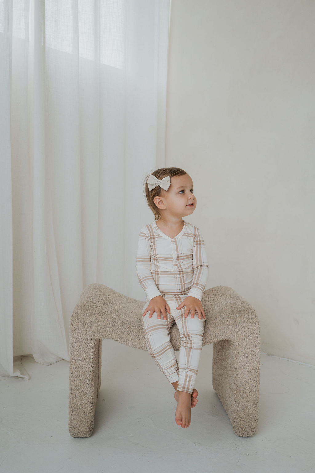A toddler wearing Bamboo Two Piece Pajamas | Ellis Plaid in a tan and white plaid two-piece set sits on a textured stool.
