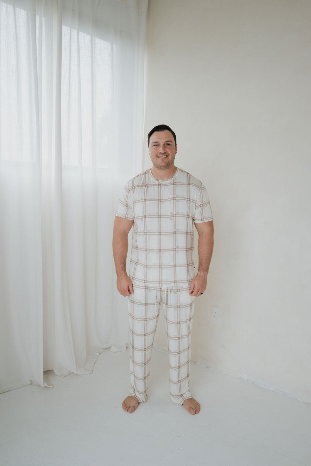 A man smiling while wearing a white and tan plaid Men’s Short Sleeve Pajamas | Ellis Plaid set consisting of a short-sleeve top and matching pants.