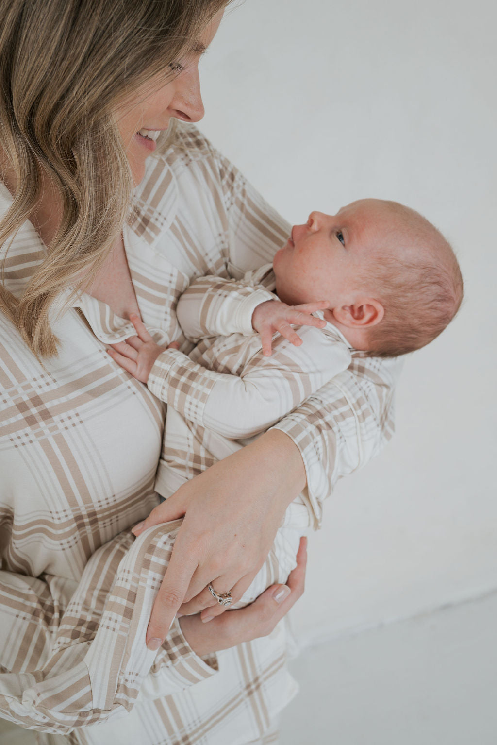 A woman holding a newborn baby, both wearing matching beige plaid Sleepy Time Set | Ellis Plaid bamboo viscose pajamas.