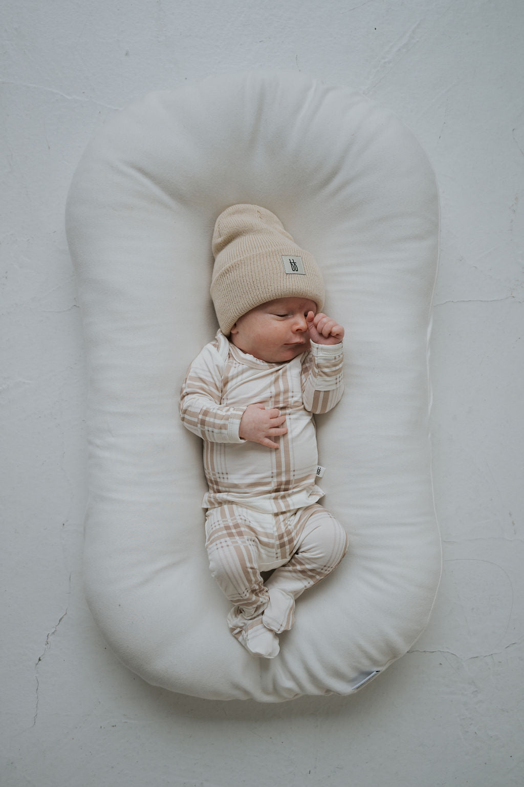 A newborn wears a Sleepy Time Set | Ellis Plaid in a beige beanie and bamboo viscose pajama sleeper while sleeping in a cream-colored lounger.