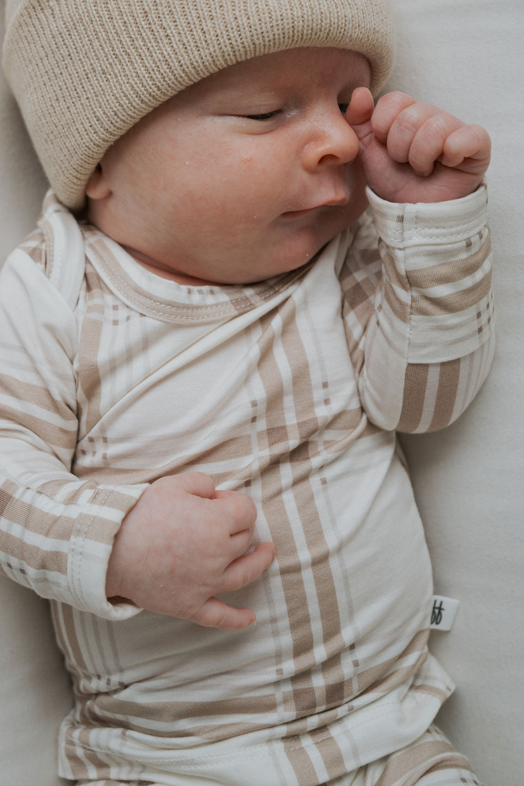 A baby wearing a tan knit beanie and the Sleepy Time Set | Ellis Plaid long-sleeved bamboo viscose bodysuit with a beige plaid pattern.