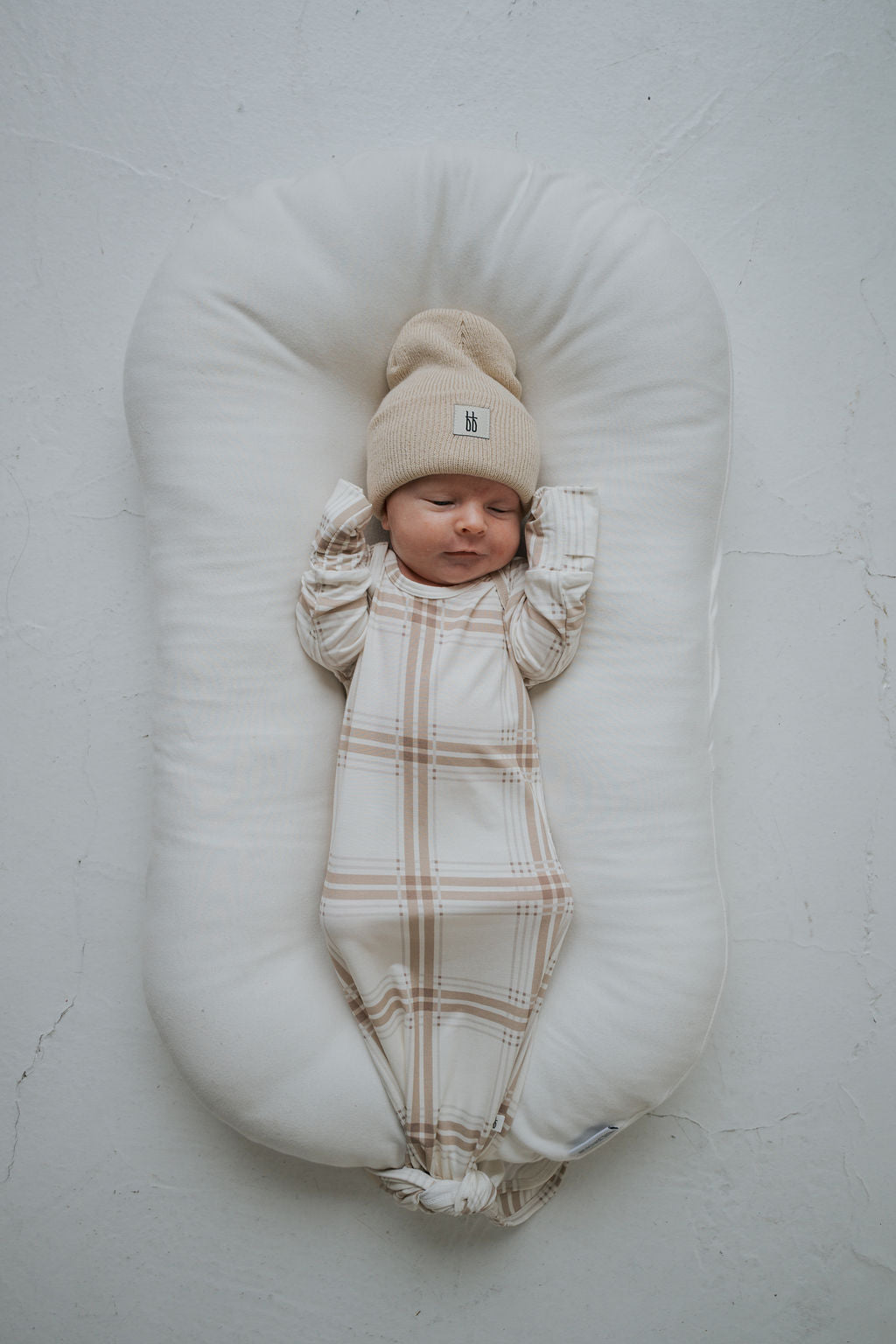 A newborn baby wearing a beige knit beanie and a Bamboo Knotted Gown | Ellis Plaid, lying in a white baby lounger.