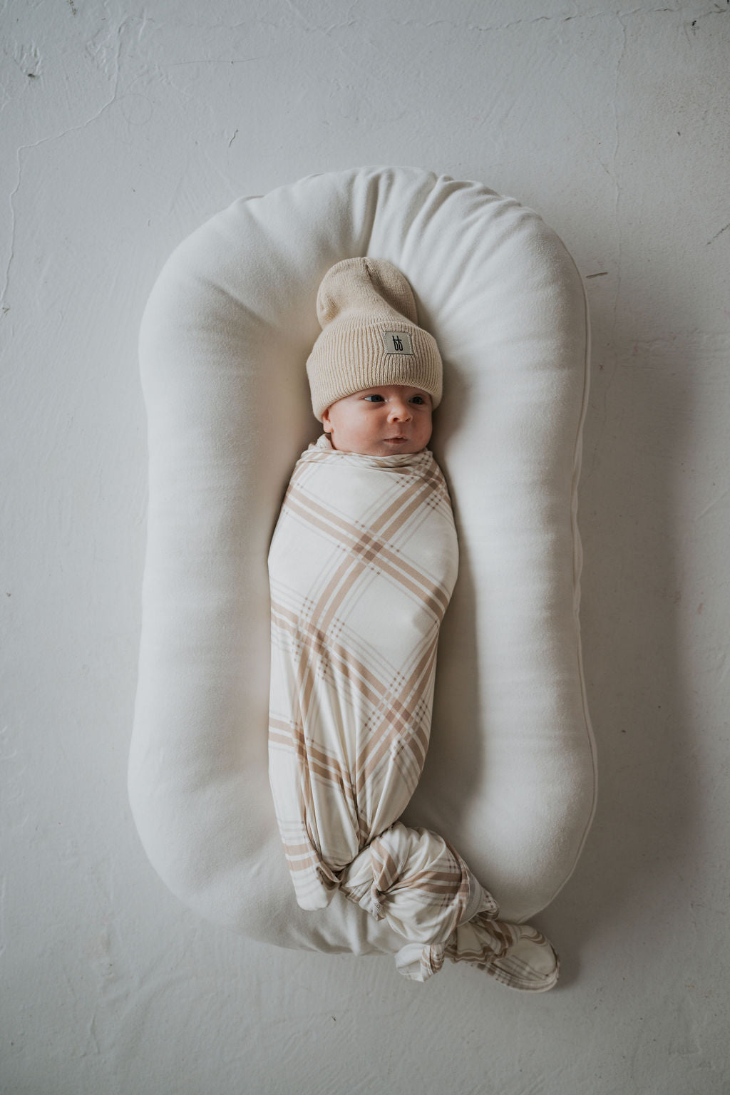An infant wearing a tan beanie lies swaddled in a Bamboo Swaddle | Ellis Plaid inside a white baby lounger.