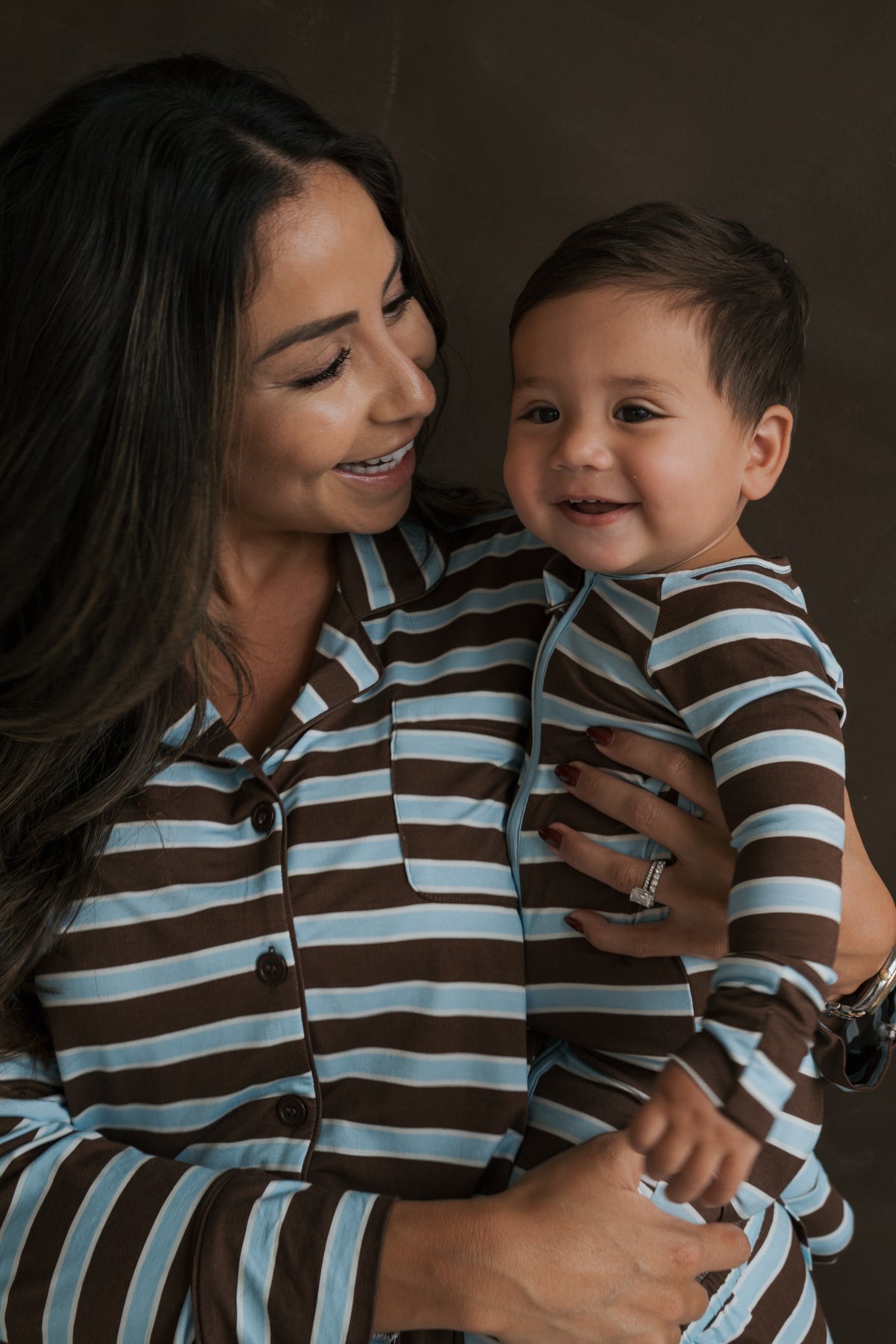 A smiling woman holding a baby, both wearing matching brown and light blue striped Bamboo Zip Pajamas | Garrett.