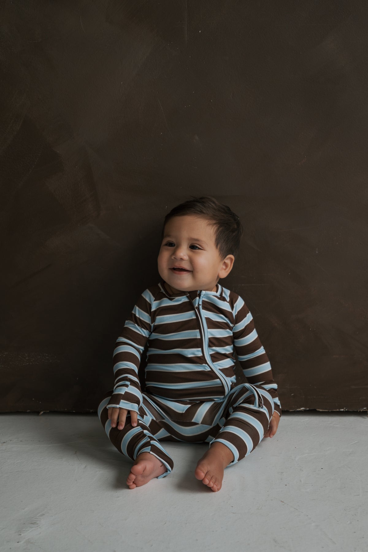 A baby sits on the floor wearing forever french baby Bamboo Zip Pajamas | Garrett, a breathable bamboo one-piece with a brown and light blue striped design and a front zipper.