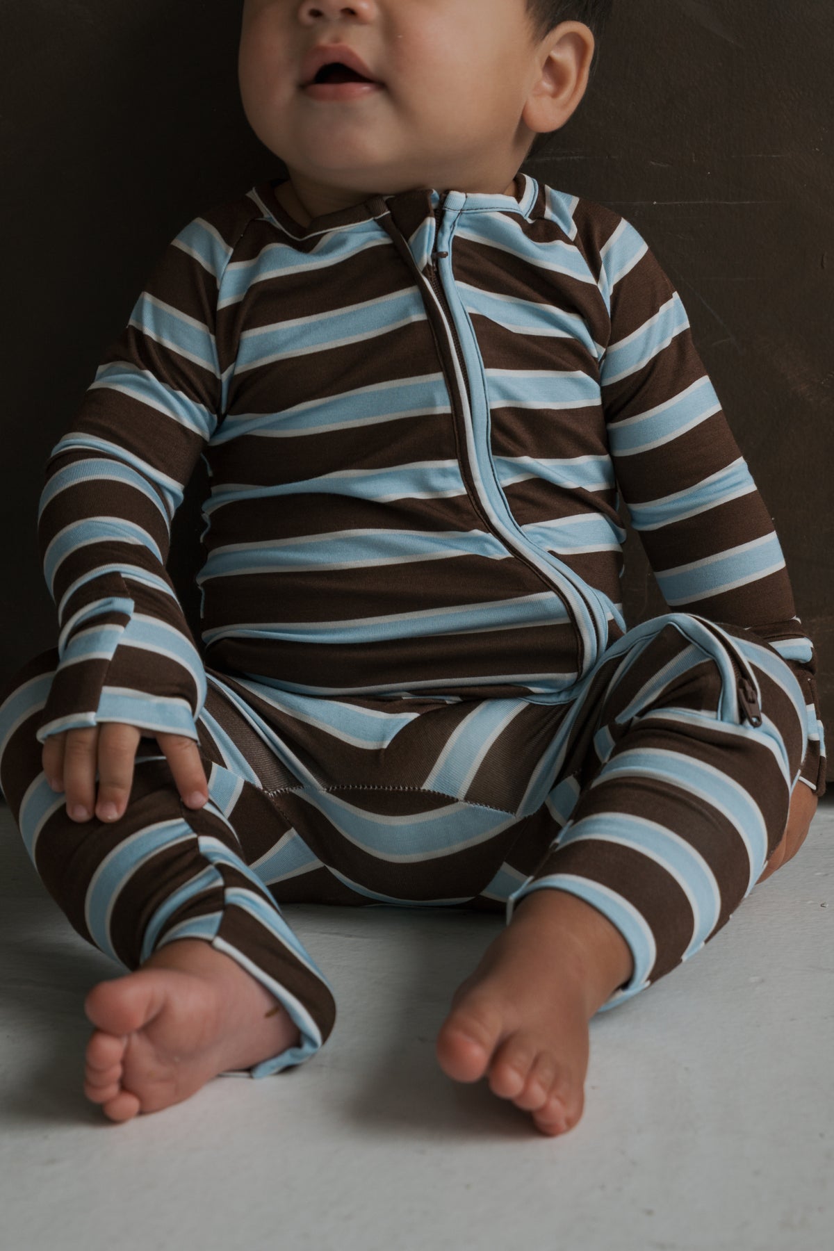 A baby wearing Bamboo Zip Pajamas | Garrett in a long-sleeved, footless, zip-up style with brown and light blue horizontal stripes.