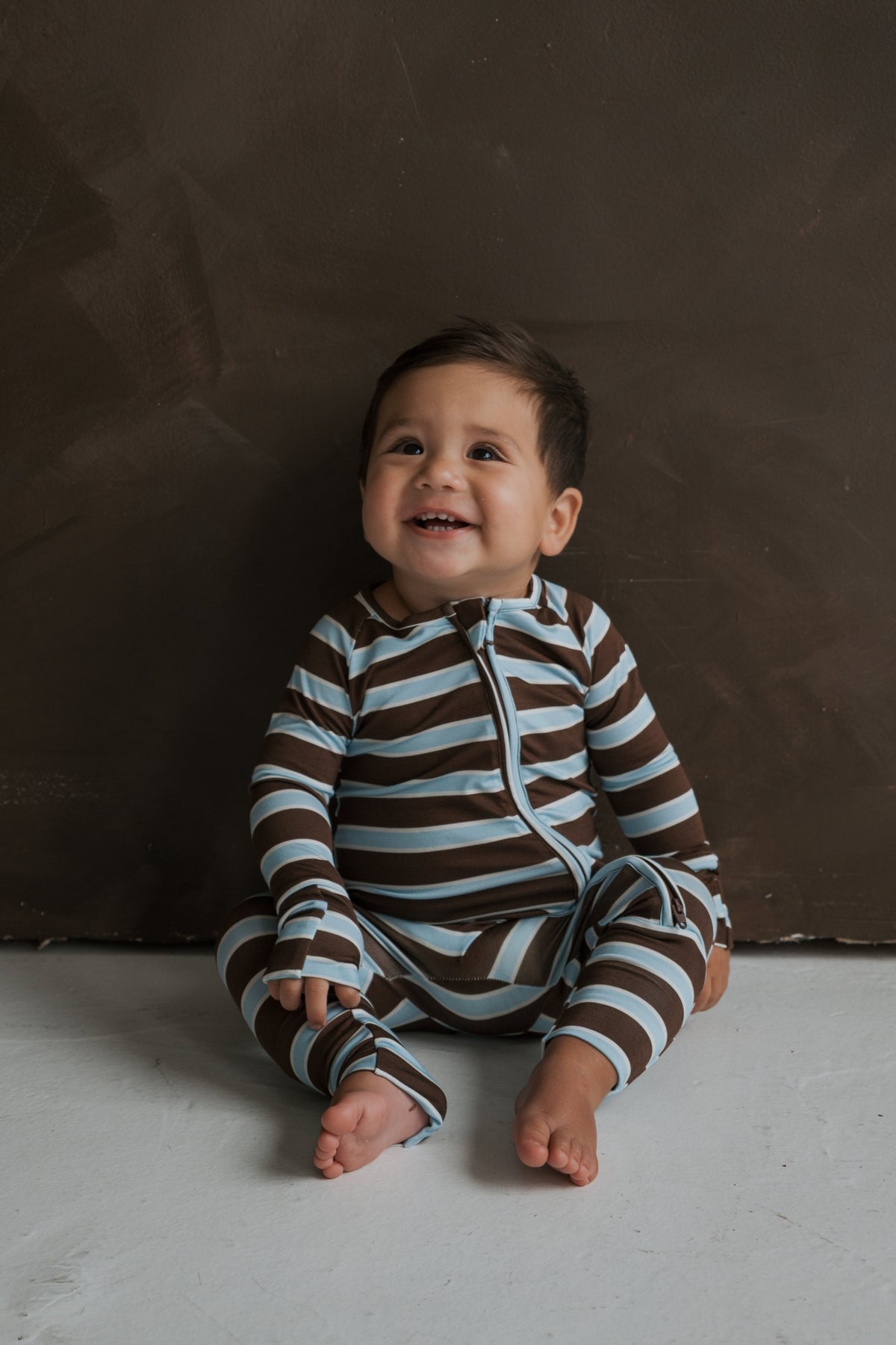 A baby smiles while wearing Bamboo Zip Pajamas | Garrett, featuring brown and light blue horizontal stripes.