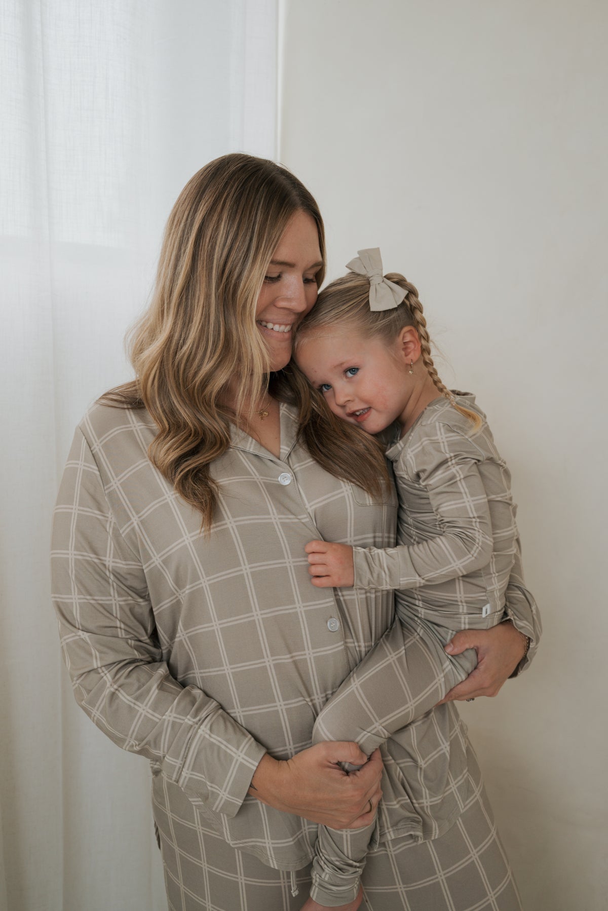 A woman holds a young child in her arms, both wearing matching Bamboo Two Piece Pajamas | Linen Grid in beige with a white grid pattern.