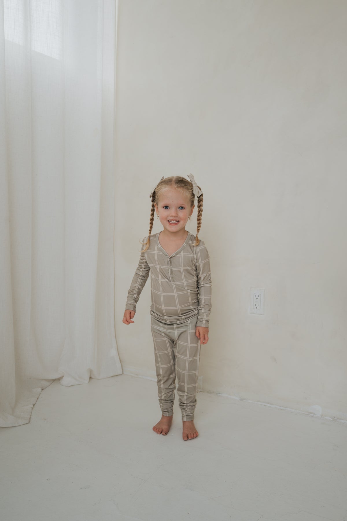 A young girl stands in forever french baby Bamboo Two Piece Pajamas | Linen Grid, featuring a beige long-sleeved shirt and matching pants with a white grid pattern.