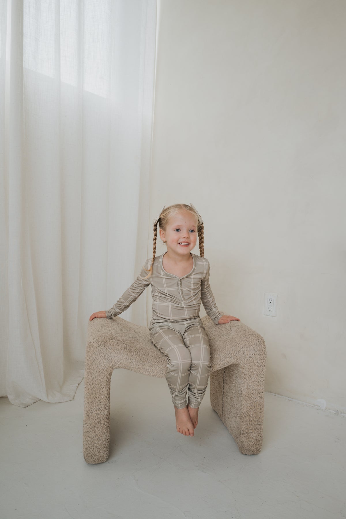 A young child with pigtails wears Bamboo Two Piece Pajamas | Linen Grid in tan with a white grid pattern while sitting on a textured, arched stool.