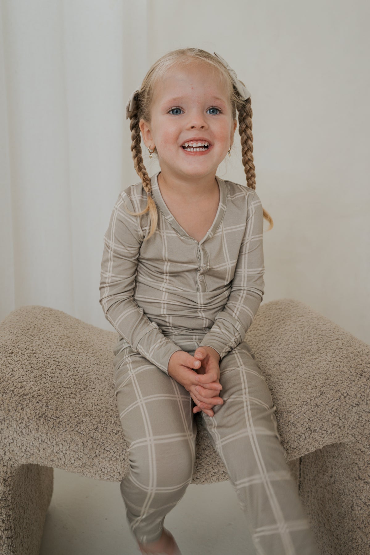 A young child smiles while wearing a beige Bamboo Two Piece Pajamas | Linen Grid set with a white grid pattern.