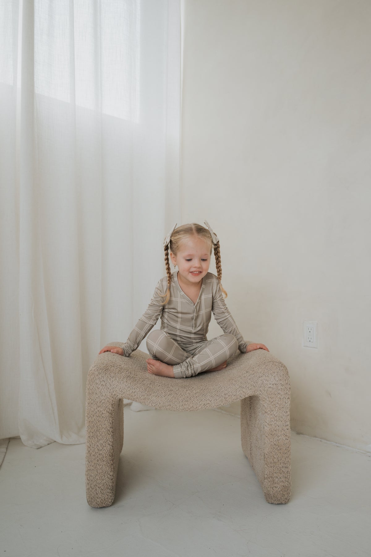 A toddler sits cross-legged on a textured stool wearing Bamboo Two Piece Pajamas | Linen Grid with a neutral grid pattern.