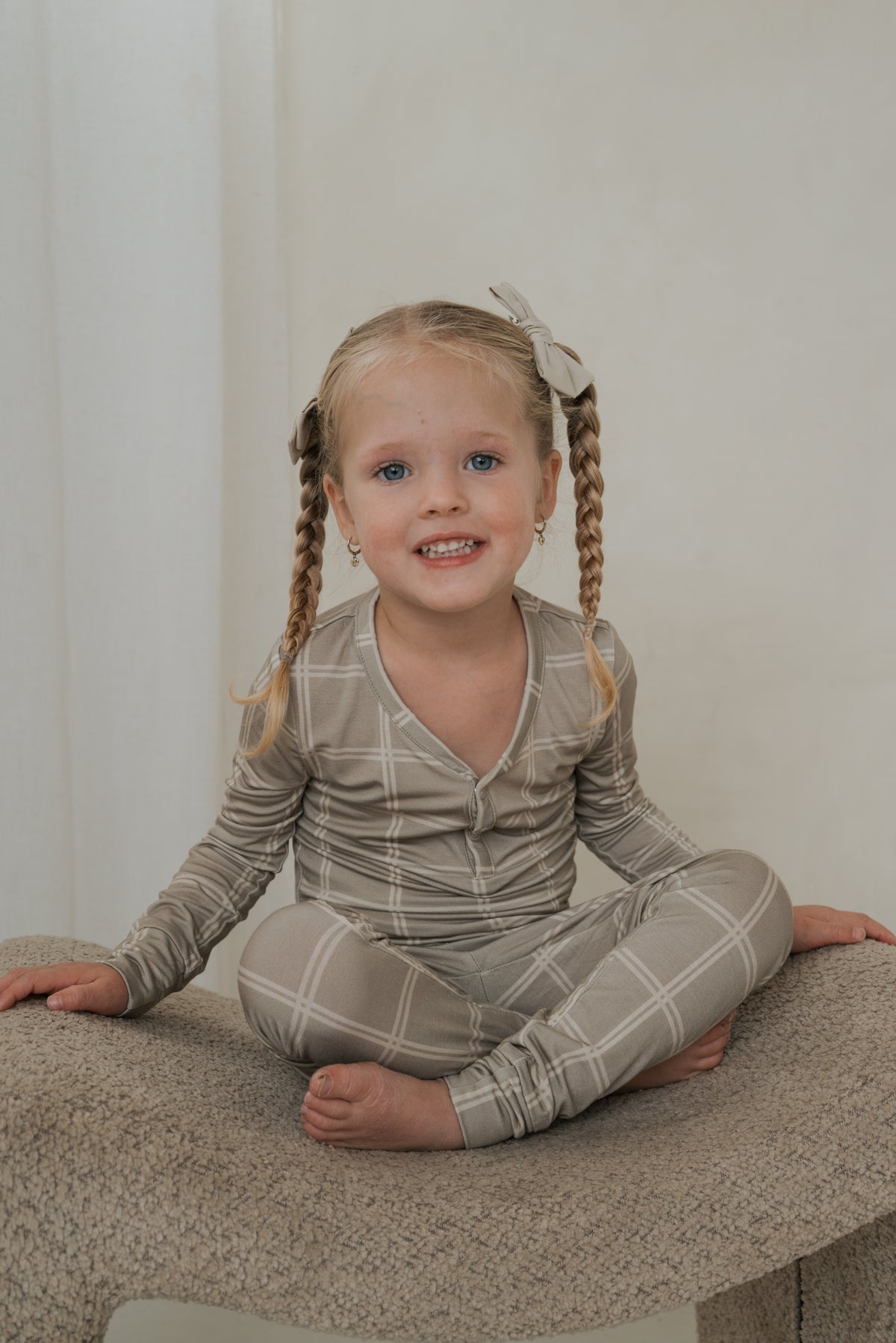 A young child sits cross-legged in Bamboo Two Piece Pajamas | Linen Grid, a long-sleeved shirt and pants set featuring a white grid pattern on a light tan background.
