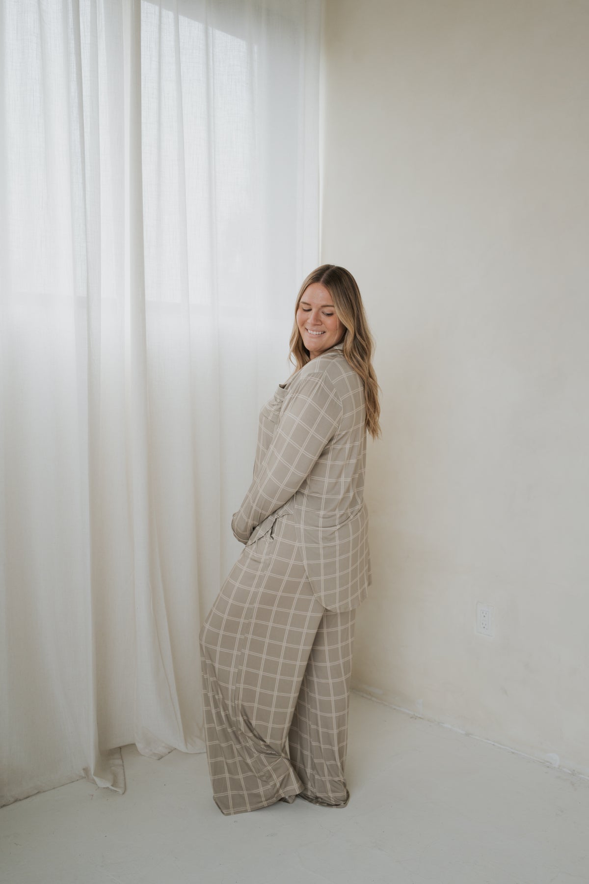 A woman wears a Women’s Bamboo Button Up Lounge Set | Linen Grid in beige, consisting of a long-sleeved button-up shirt and matching wide-leg trousers, both featuring a white grid pattern.