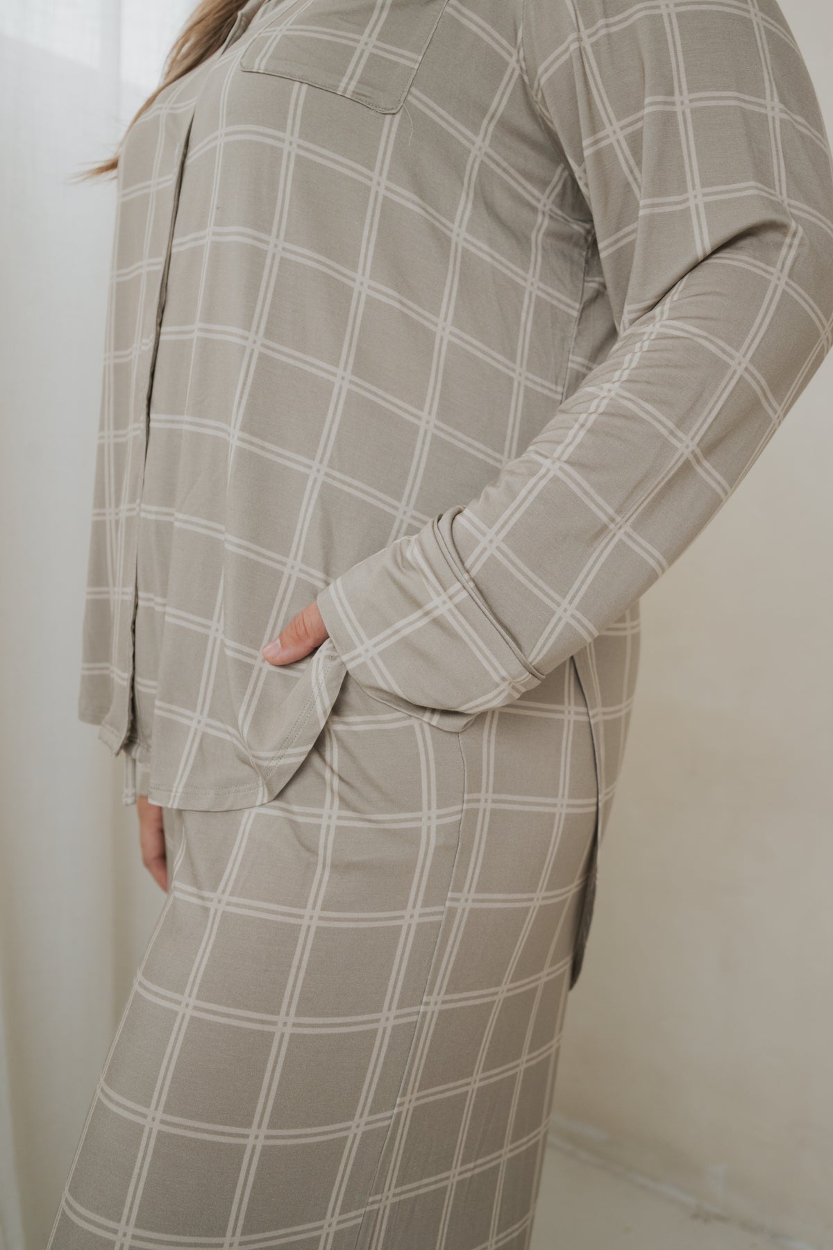 A person wears a beige grid-patterned Women’s Bamboo Button Up Lounge Set | Linen Grid, consisting of a long-sleeved button-up shirt and wide-leg lounge pants, with their hand in the shirt’s front pocket.