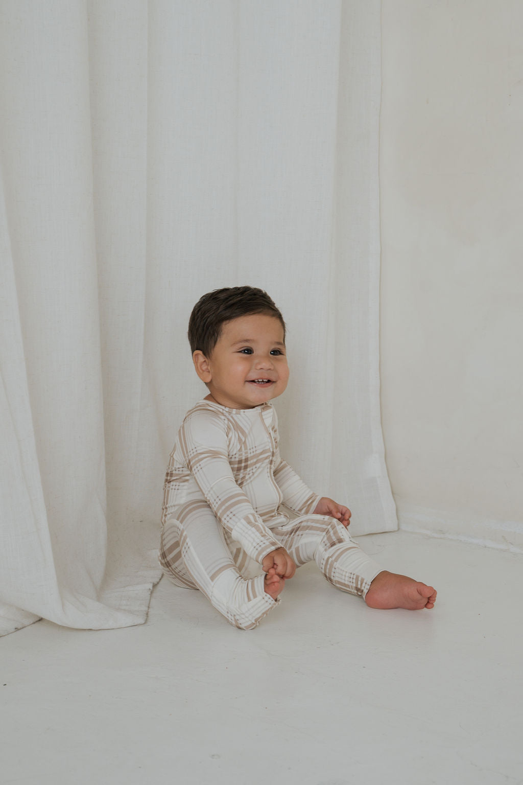 A smiling baby sits on the floor, wearing a tan plaid Bamboo Zip Pajamas | Ellis Plaid bamboo viscose sleeper.