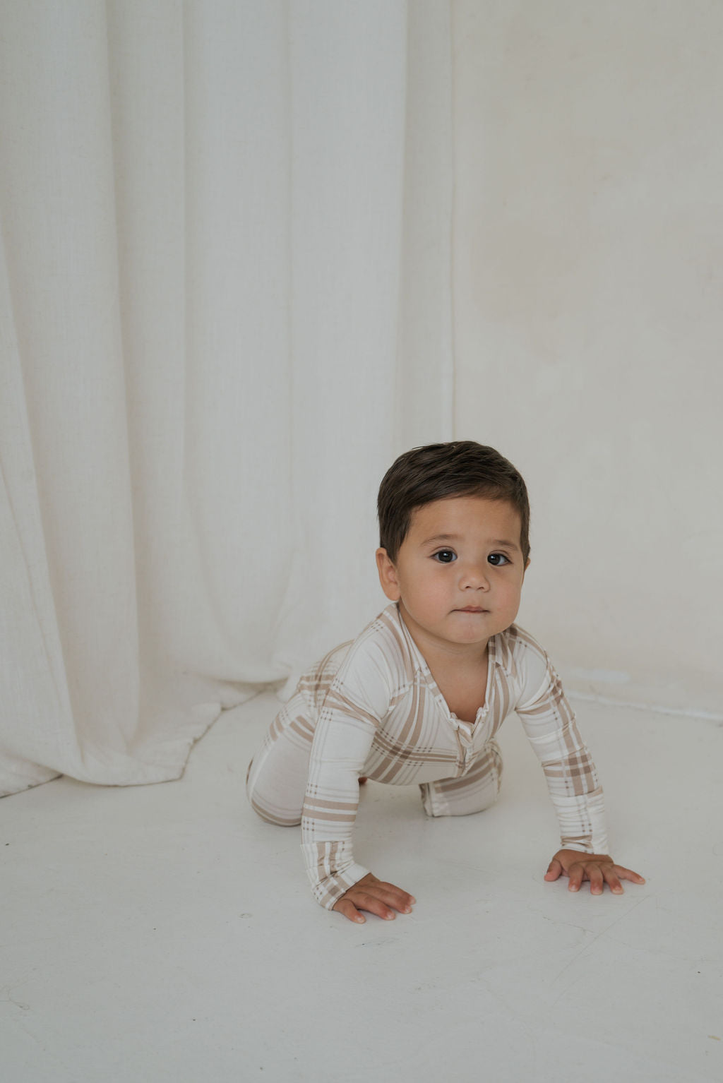 A toddler crawls on the floor wearing a cream-colored Bamboo Zip Pajamas | Ellis Plaid bamboo viscose zip-up sleeper with a brown plaid pattern.