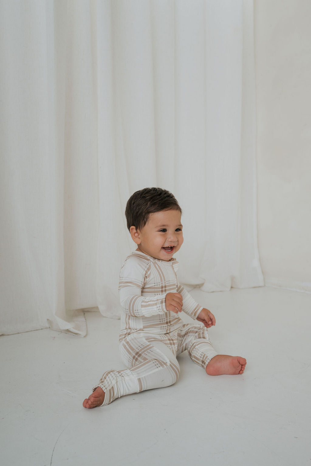 A laughing baby sits on the floor wearing a long-sleeved, footed Bamboo Zip Pajamas | Ellis Plaid in a tan and white plaid pattern.
