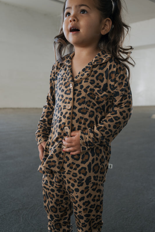 A child wearing a brown and black Girl’s Bamboo Button Up Lounge Set | Leopard, consisting of a long-sleeved button-up shirt and matching pants.