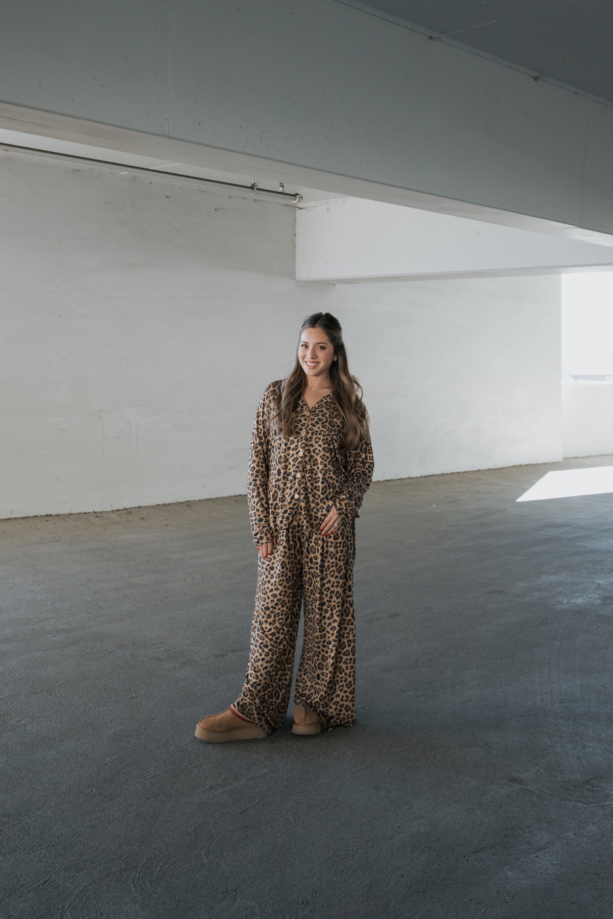 A smiling woman stands in a parking garage wearing a forever french baby Women’s Bamboo Button Up Lounge Set in leopard print and platform slippers.