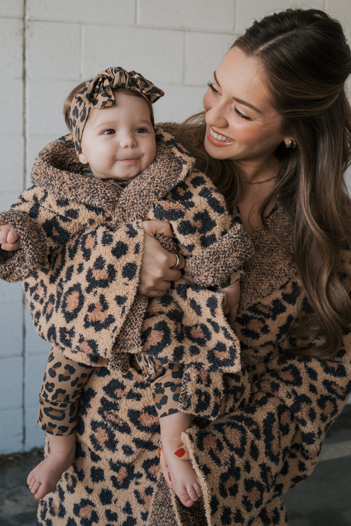 A woman holds a baby wearing a forever french baby Children’s Robe | Leopard, an Oeko-tex certified, leopard-print hooded baby robe with a matching headband.