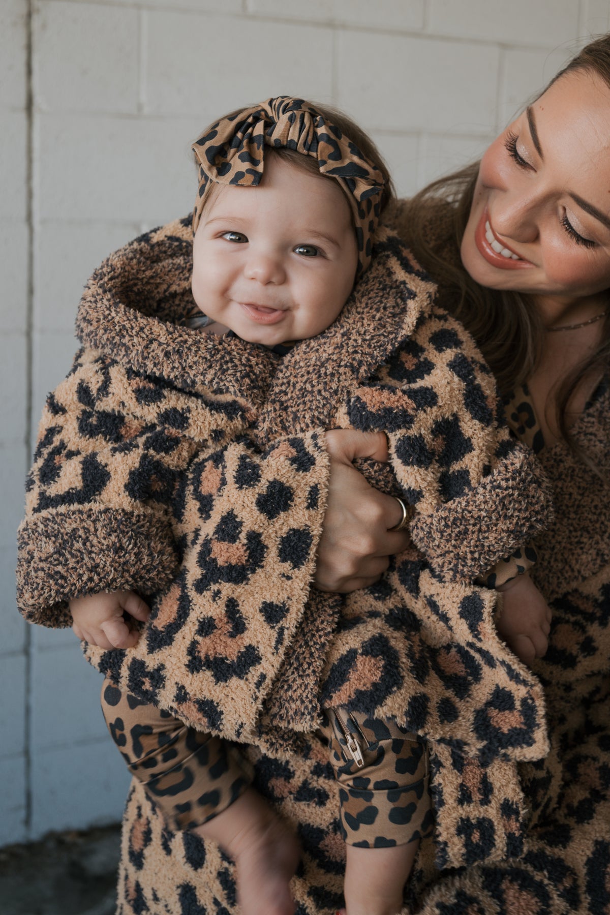 A woman holds a smiling baby wearing an Oeko-tex certified, hooded Children’s Robe | Leopard and matching headband.
