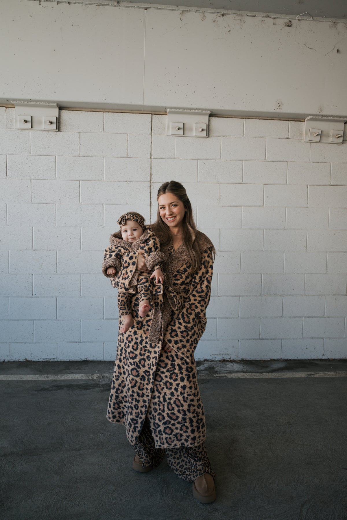 A woman smiling while holding an infant, both wearing matching Adult Robe | Leopard robes and leopard print headwear.