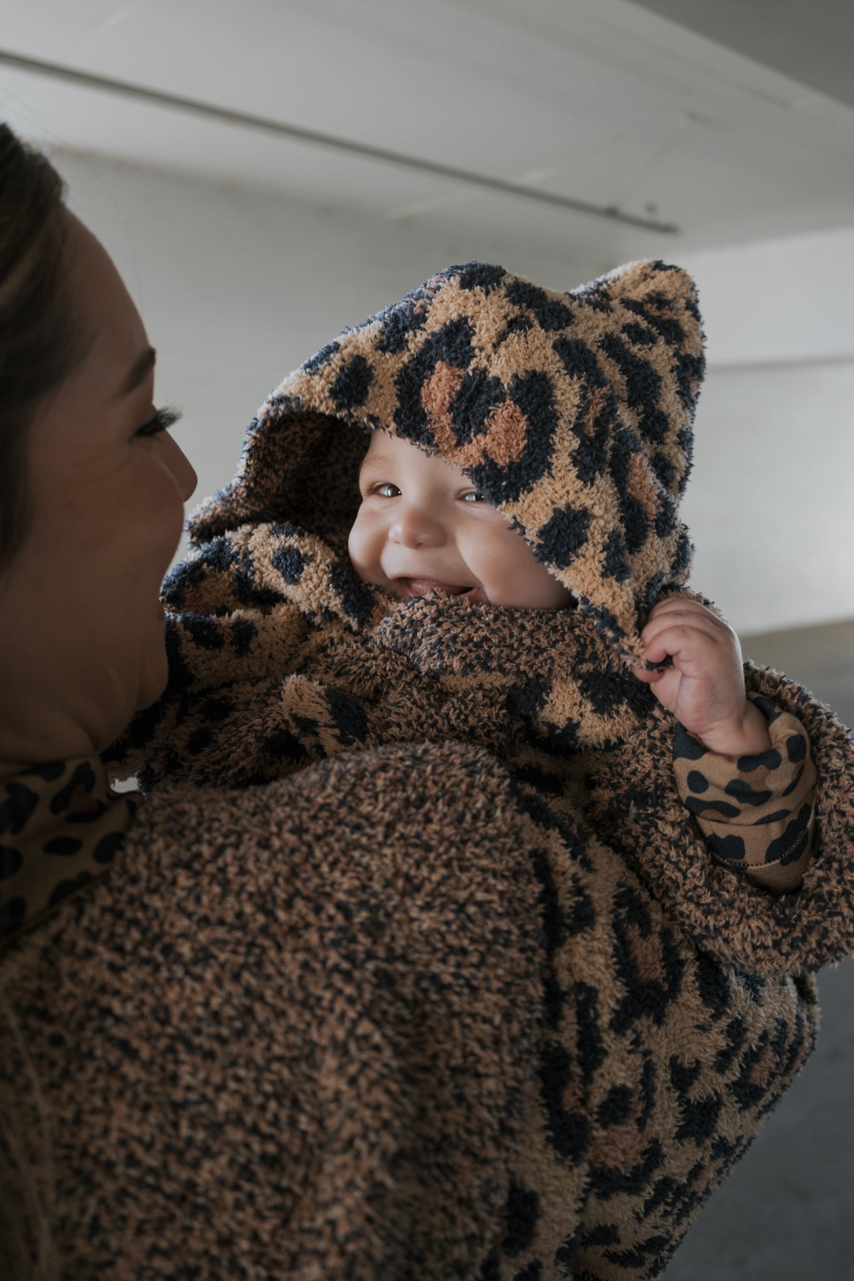 A smiling baby wearing a plush, Oeko-tex certified Children’s Robe | Leopard hood held by a woman.