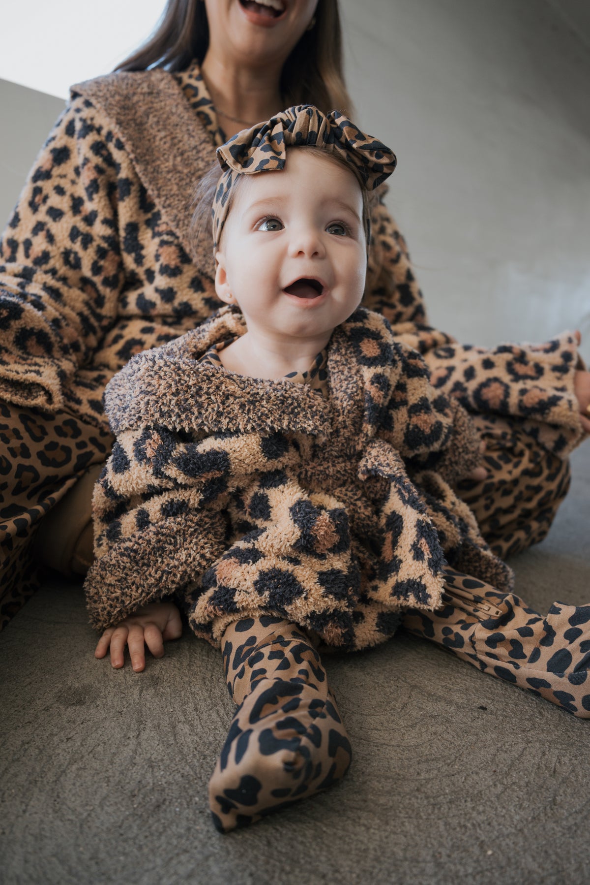 A baby wearing an Oeko-tex certified Children’s Robe | Leopard with a matching headband sits on the floor in front of an adult wearing a matching leopard print robe.