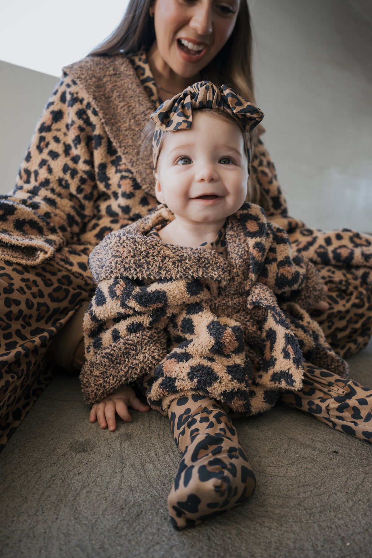 A baby wearing an Oeko-tex certified Children’s Robe | Leopard with matching headband, seated in front of an adult wearing a matching robe.