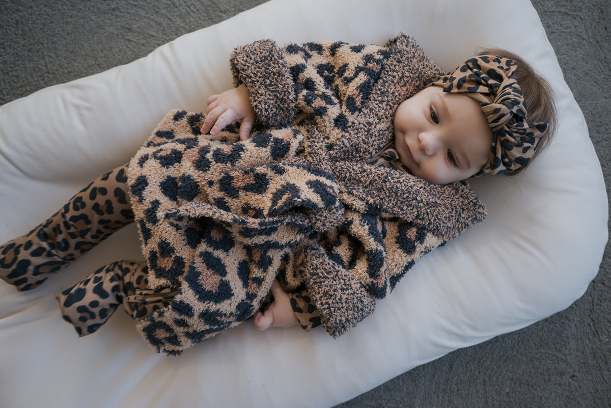 A baby wearing a Children’s Robe | Leopard with a hood, matching leggings, and a knotted headband while lying on a cushioned surface.