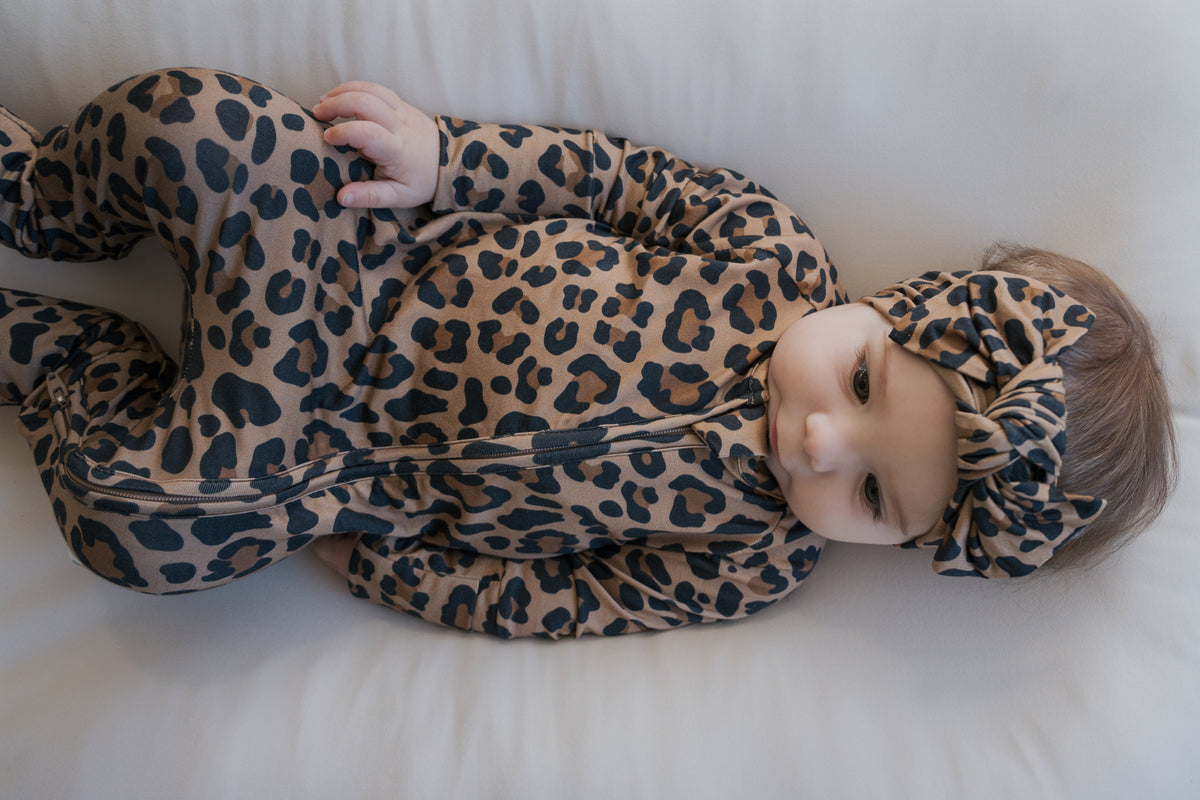 A baby wearing leopard print Bamboo Zip Pajamas | Leopard with a zip-up footie design and a matching knotted headband.