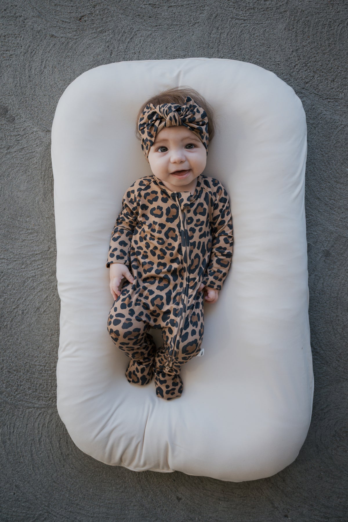 A baby wearing Leopard Bamboo Zip Pajamas and a matching knotted headband, lying in a cream-colored lounger.