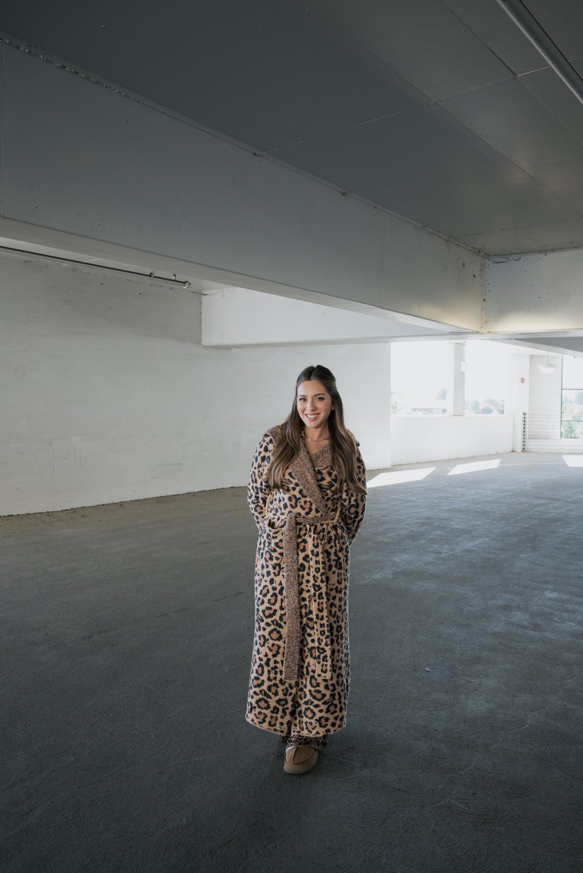 A woman wears a full-length leopard print Adult Robe | Leopard by forever french baby while standing in an open parking garage.