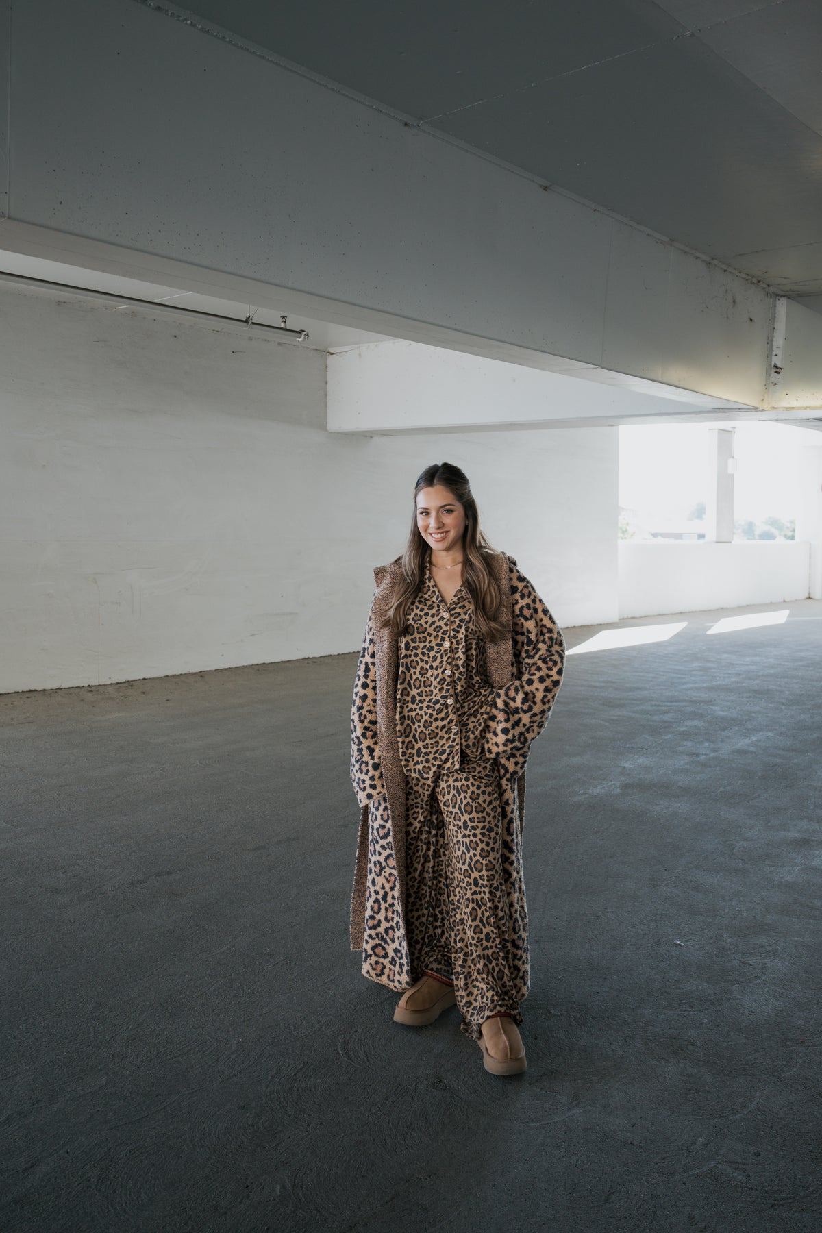 A smiling woman wears an Adult Robe | Leopard by forever french baby over a matching leopard-print shirt and pants, paired with tan platform shoes.