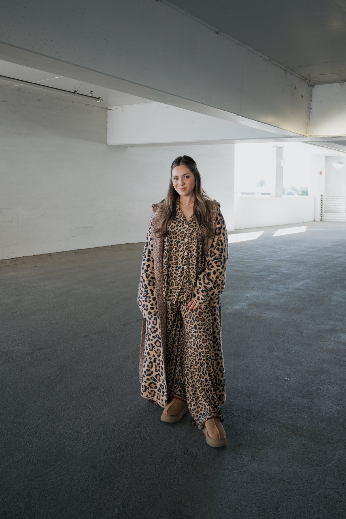 A person wearing a forever french baby Adult Robe | Leopard over a matching shirt and pants with platform clogs stands in a parking garage.